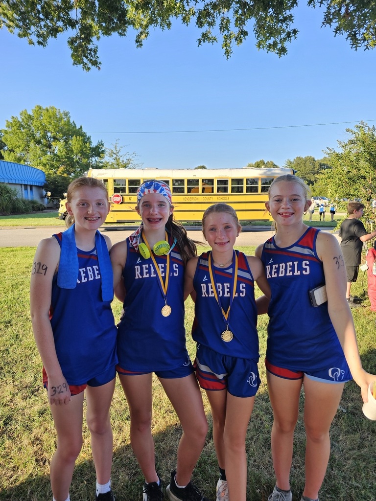Four young Obion County Rebels cross country runners pose together on a grassy field, wearing matching blue-and-red uniforms. Two runners wear medals, and one wears a colorful headband and headphones. A yellow school bus is visible in the background along with trees, a school building, and other runners and spectators.
