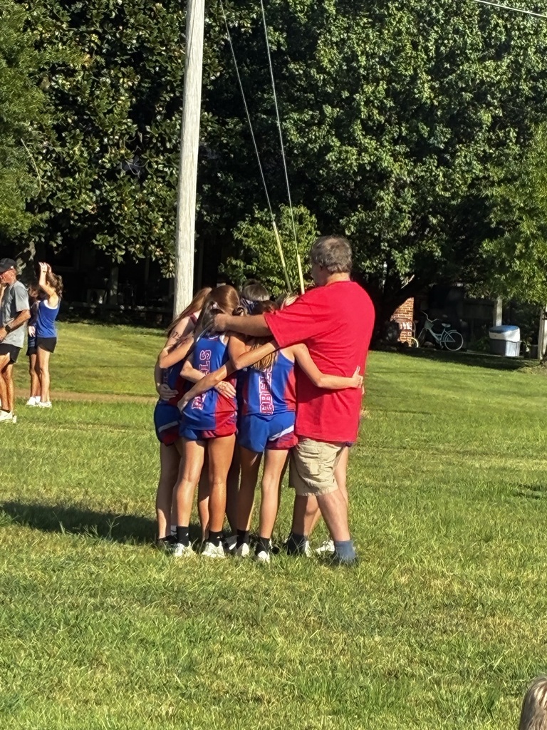 A group of young athletes in blue and red uniforms huddle together on a grassy field with their coach, who wears a red shirt and khaki shorts. The group stands closely with arms around one another in a team moment. Trees, spectators, a utility pole, bicycles, and a trash can are visible in the background.