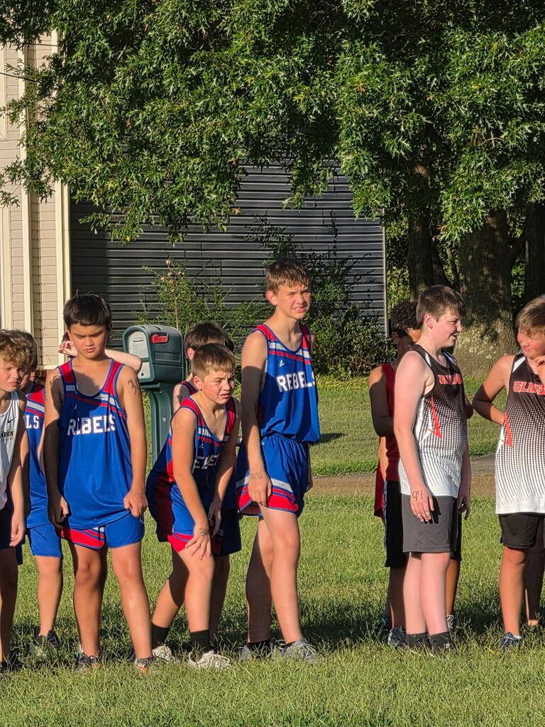 Young cross country runners stand together on a grassy field before or after a race. Several athletes wear blue-and-red Rebels uniforms, while others wear black, gray, and red team uniforms. Trees and a building are visible in the background.