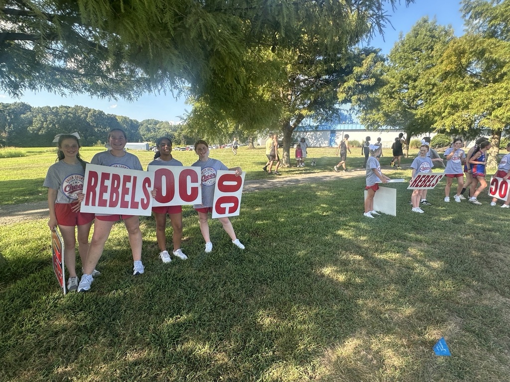Obion County Middle School cheerleaders stand on a grassy field in the shade of trees, smiling and holding large red-and-white signs that read “REBELS,” “OC,” and “GO.” Other cheerleaders pose with signs in the background, while students, spectators, a school building, and parked vehicles are visible across the field.