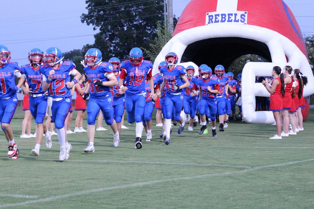 A youth football team in blue and red uniforms runs onto the field through a large inflatable football helmet labeled “REBELS,” while cheerleaders in red uniforms line both sides of the entrance.
