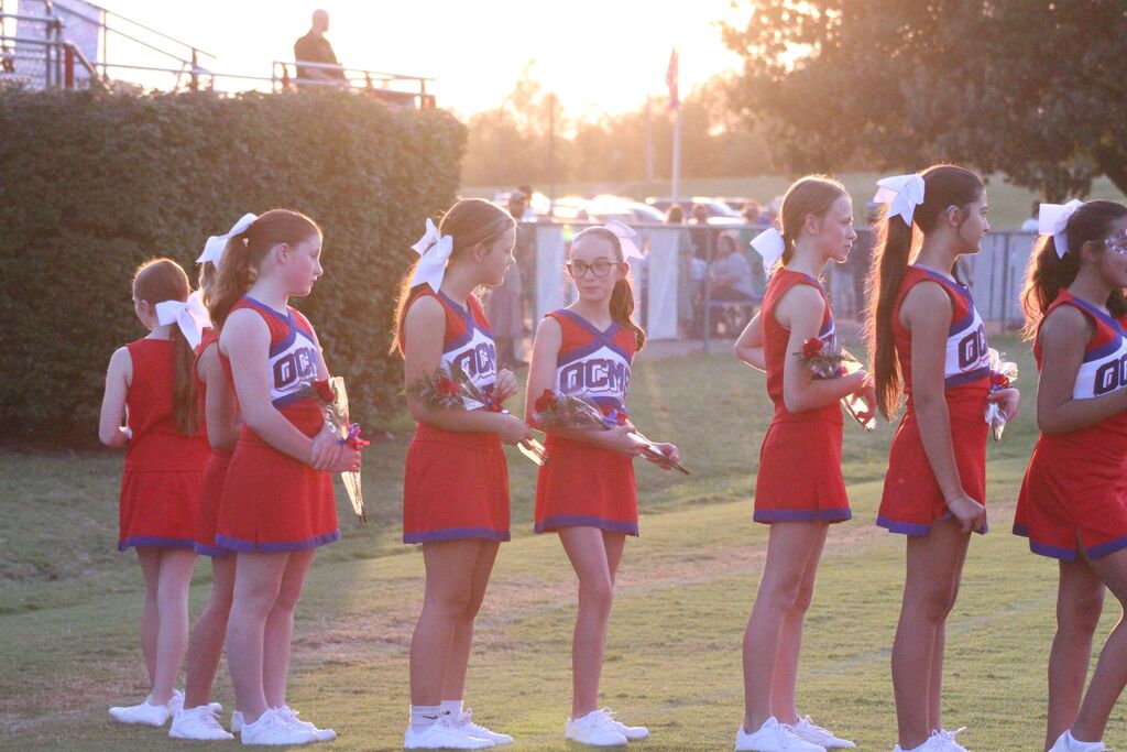 OCMS cheerleaders in red, white, and blue uniforms stand in a line on the football field, holding small bouquets of flowers. Large white bows adorn their hair as they participate in a sunset ceremony or recognition event.