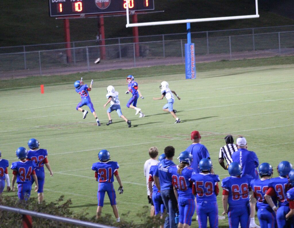A youth football player in a blue and red uniform leaps to catch a pass while two defenders in white uniforms close in. Teammates and coaches watch from the sideline, with the goalpost and scoreboard visible in the background.