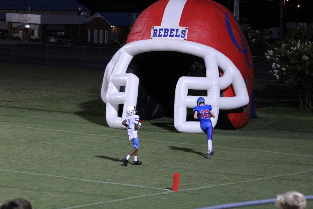 A football player in a blue uniform runs toward the end zone with a defender in a white uniform nearby. Behind them is a large inflatable red football helmet marked “REBELS,” while the end-zone pylon and field are visible in the foreground.