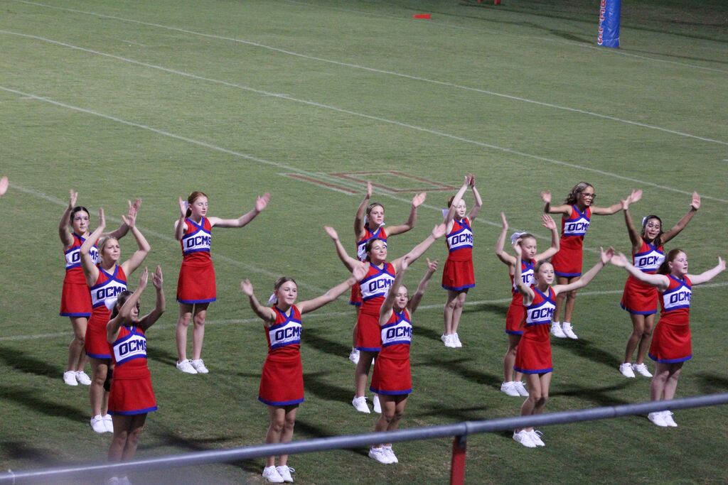 A group of OCMS cheerleaders in red, white, and blue uniforms perform a routine on the football field, raising their arms in synchronized motions during a game.