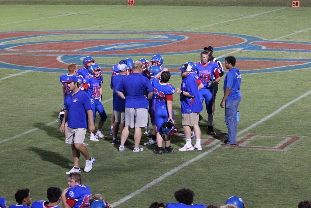 A youth football team in blue and red uniforms gathers with coaches on the field during a game. Players stand in a huddle near midfield, with the large painted field logo, yard lines, and a referee visible in the background.