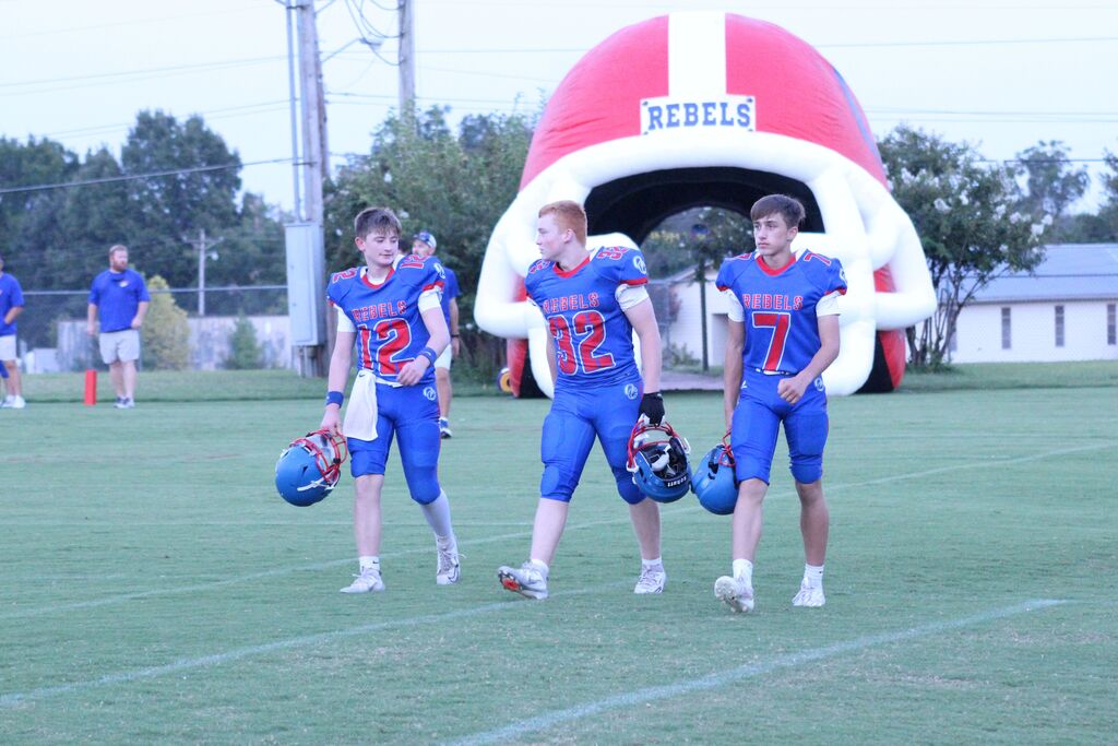Three youth football players in blue and red “Rebels” uniforms walk across the field carrying their helmets, with a large inflatable football helmet entrance behind them.