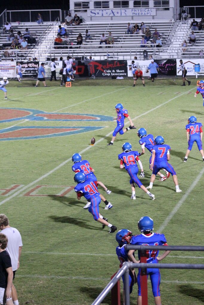 Youth football players in blue and red uniforms line up for a play as one player prepares to kick the football. Teammates move downfield while coaches, officials, and spectators watch from the sideline and bleachers.