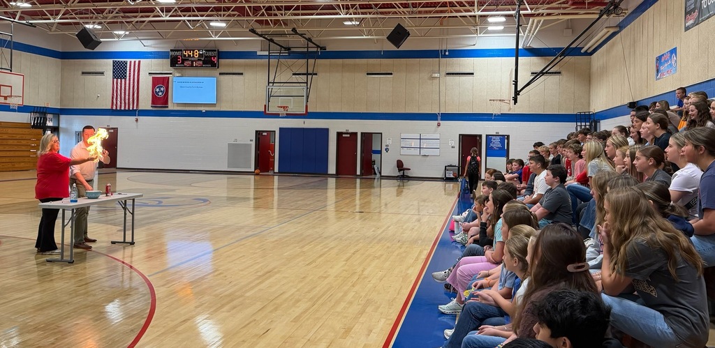 Students sit in the OCMS gym bleachers watching an FCA demonstration. At the front of the gym, Mrs. Angie Partin participates in a controlled fire demonstration alongside Brother Eric as part of a lesson about Shadrach, Meshach, and Abednego.
