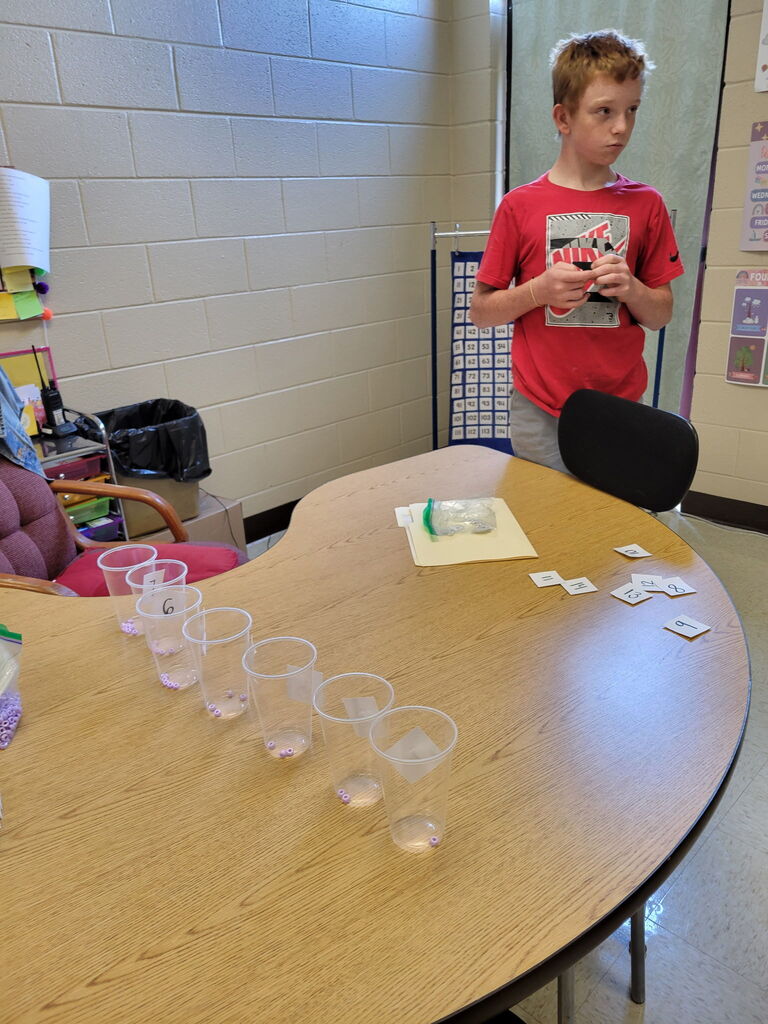 Carson Butler stands beside a classroom table while working on a hands-on math activity using numbered cups, beads, and number cards.