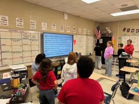Students gather near the classroom display to read a scenario and collaborate on possible solutions.