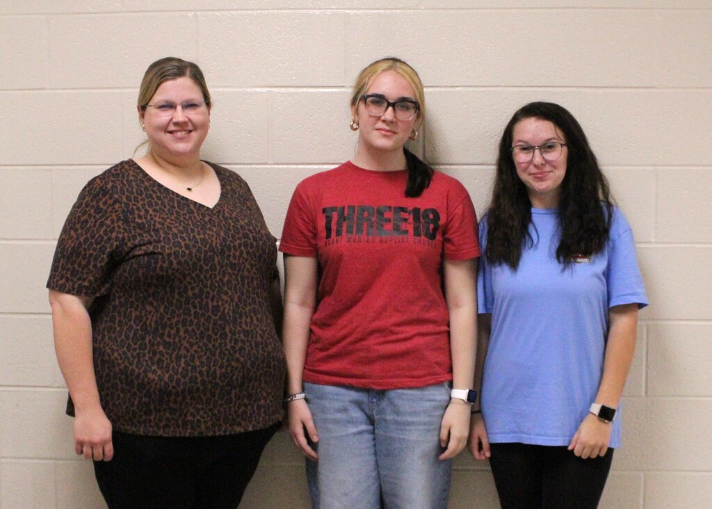 Three women pose for a photo in front of a beige brick wall. The woman on the left wears a leopard-print top and black pants, while the two young women beside her wear glasses; the center woman wears a red T-shirt and jeans, and the woman on the right wears a light blue shirt and black pants.