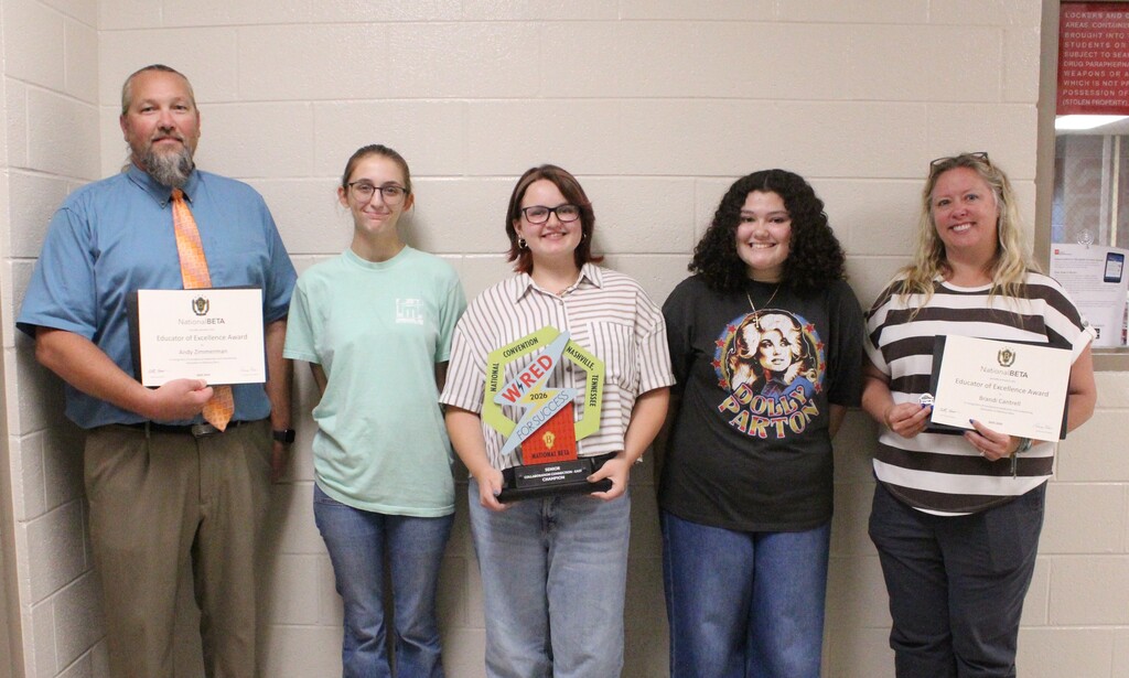 Five people pose for a photo in front of a beige brick wall. Two adults hold National Beta Educator of Excellence Awards, while three students stand between them. The center student holds a National Beta 2026 convention trophy.