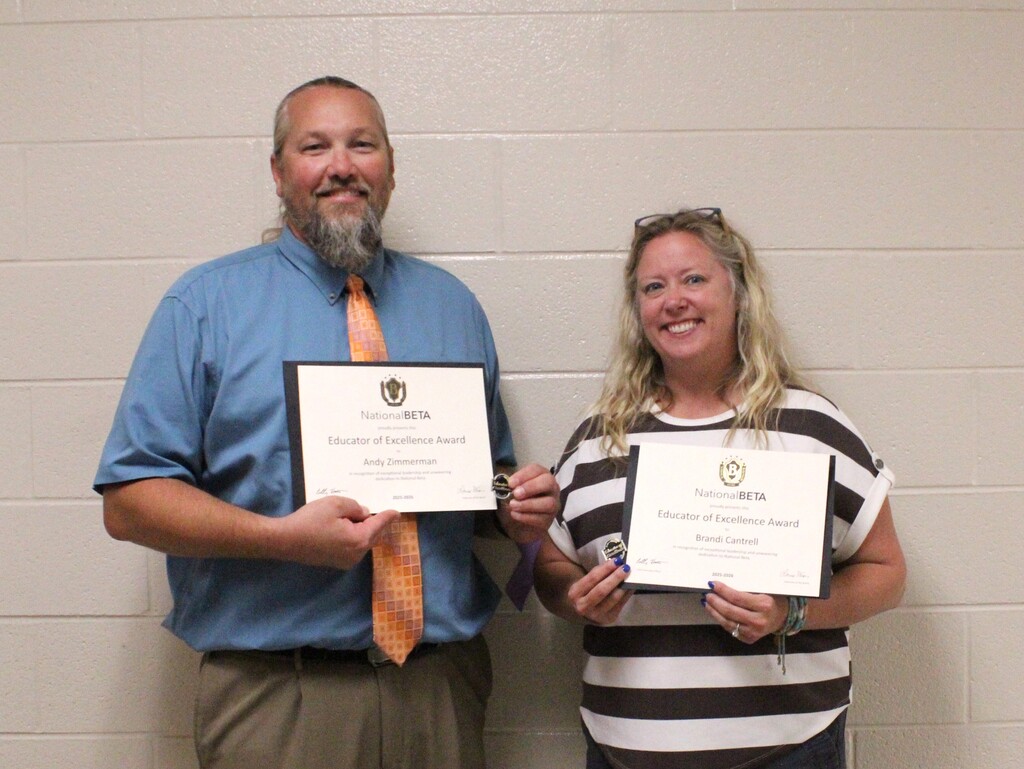 Two adults pose in front of a beige brick wall, each holding a National Beta Educator of Excellence Award certificate and medal. The man on the left wears a blue shirt and orange tie, while the woman on the right wears a black-and-white striped top. Both are smiling at the camera.