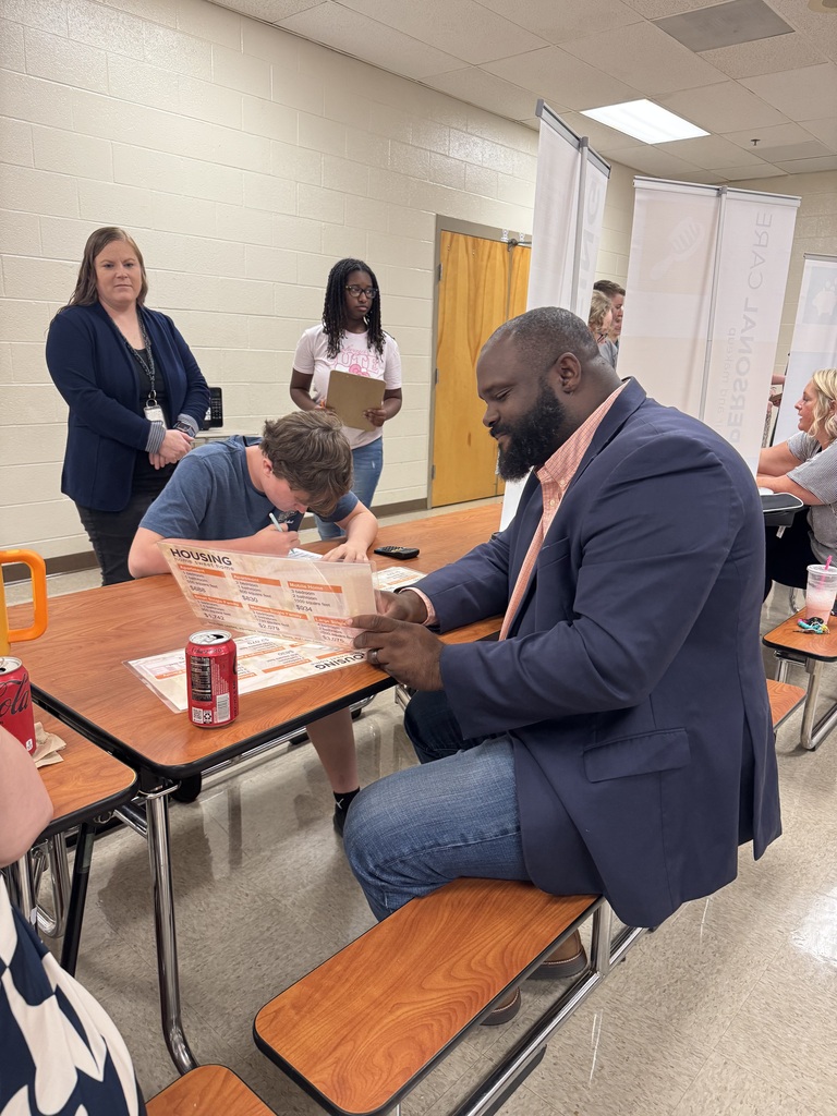 A South Fulton Middle School student works through a housing budget with an adult facilitator during the school’s Financial Fair. The student completes a budgeting worksheet while reviewing housing options and expenses. Other students and facilitators participate in the hands-on financial literacy activity in the background.