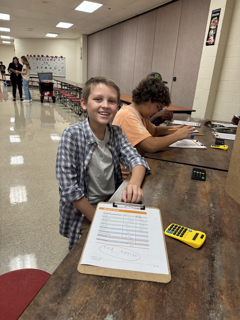 A South Fulton Middle School student smiles while completing a budgeting activity during the school’s Financial Fair. The student works with a budget sheet and calculator as other students complete similar financial exercises at nearby stations.