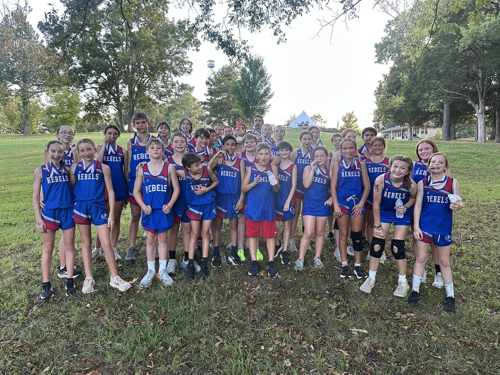 A large group of young cross country runners poses together outdoors on a grassy hill after a meet, wearing matching blue and red Rebels uniforms and several displaying race medals.