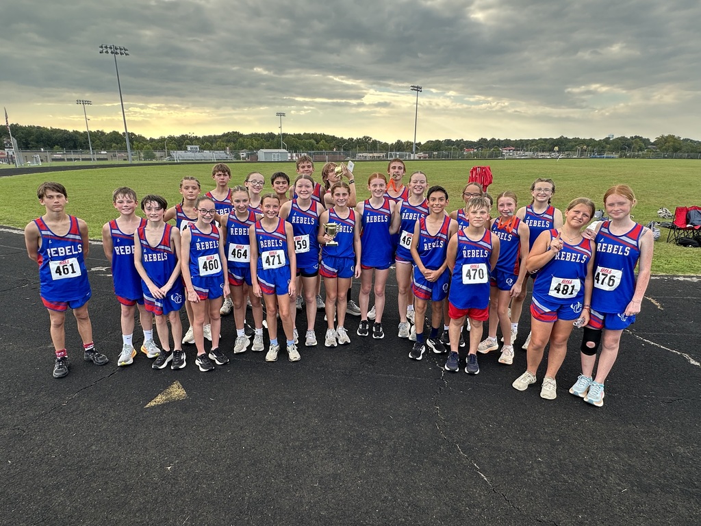 A group of young cross country runners in matching blue and red Rebels uniforms poses on a track beside a large grassy athletic field. Several runners wear race bibs, while others hold medals, a trophy and awards.