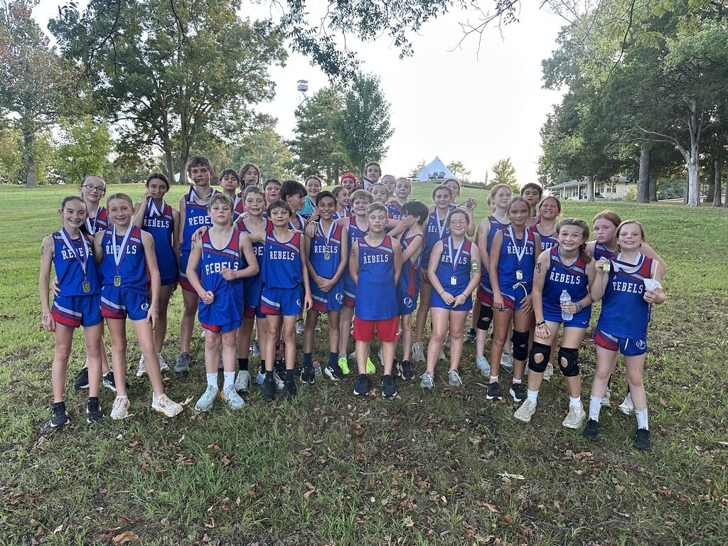 A large group of young cross country runners poses together on a grassy hill after a meet, wearing matching blue and red Rebels uniforms. Several runners are wearing medals, while others hold water bottles and snacks.