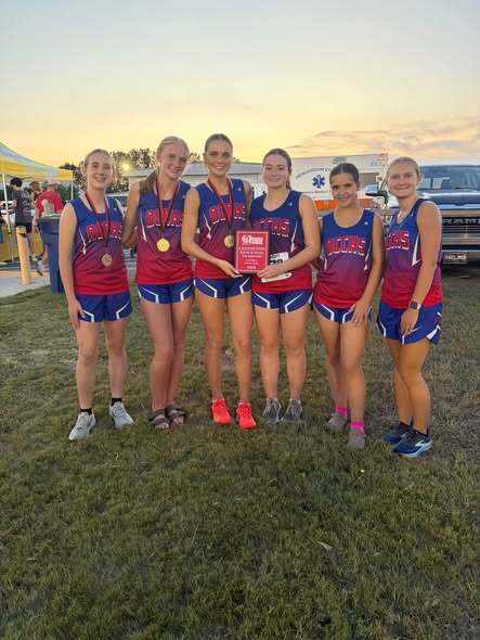 Six girls from OCCHS cross country stand together outdoors after a meet, wearing matching red and blue uniforms and medals. Two girls hold a first-place team trophy and a red award plaque.