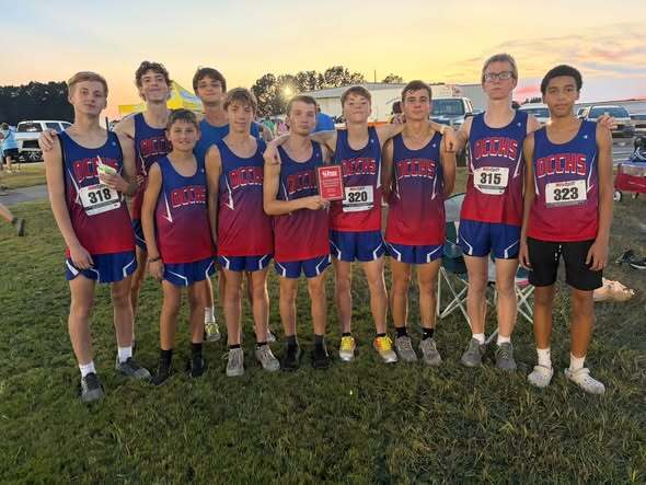 Nine OCCHS boys cross country runners pose together outdoors after a meet, wearing matching red and blue uniforms. Several runners wear race bibs, and two runners hold a red award plaque.