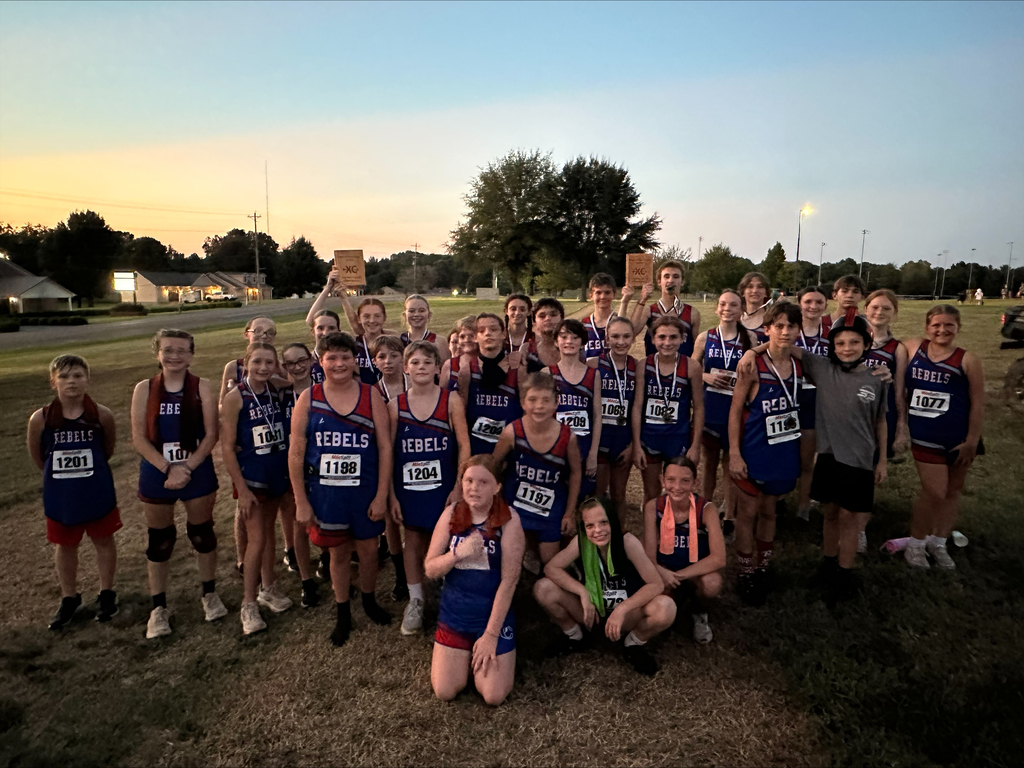 Group photo of Obion County Middle School cross-country runners in blue and red “Rebels” uniforms gathered outdoors after the Best of the West meet. Several runners hold wooden cross-country awards, and some wear medals around their necks.