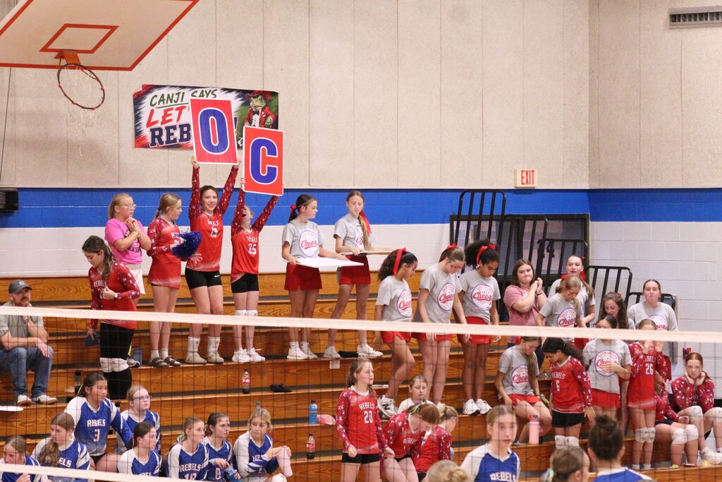 Cheerleaders and JV Volleyball players cheer in the stands and hold up signs spelling out "OC" during volleyball game.