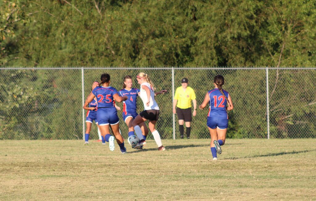Girls soccer players in blue and white uniforms compete for the ball on a grassy field during a game, with a referee watching the play in the background.