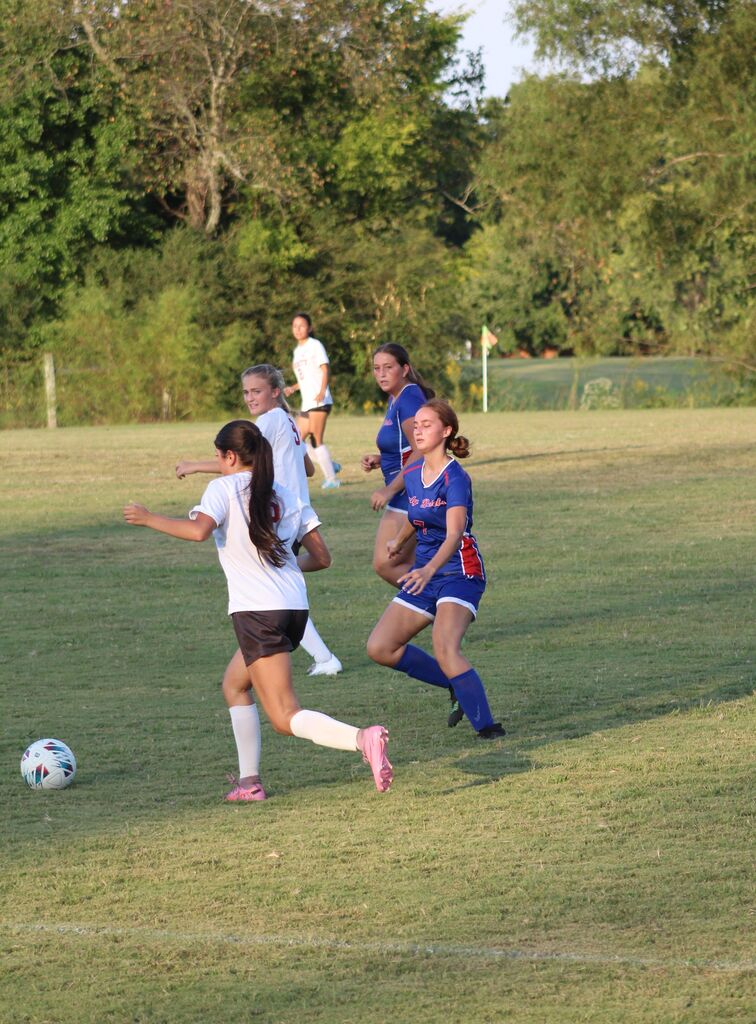 Girls soccer players in blue and white uniforms run toward the ball during a game on a grassy field, with trees and a flag in the background.