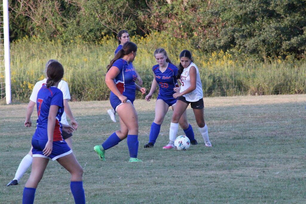 Girls soccer players in blue and white uniforms compete for possession of the ball on a grassy field during a game.