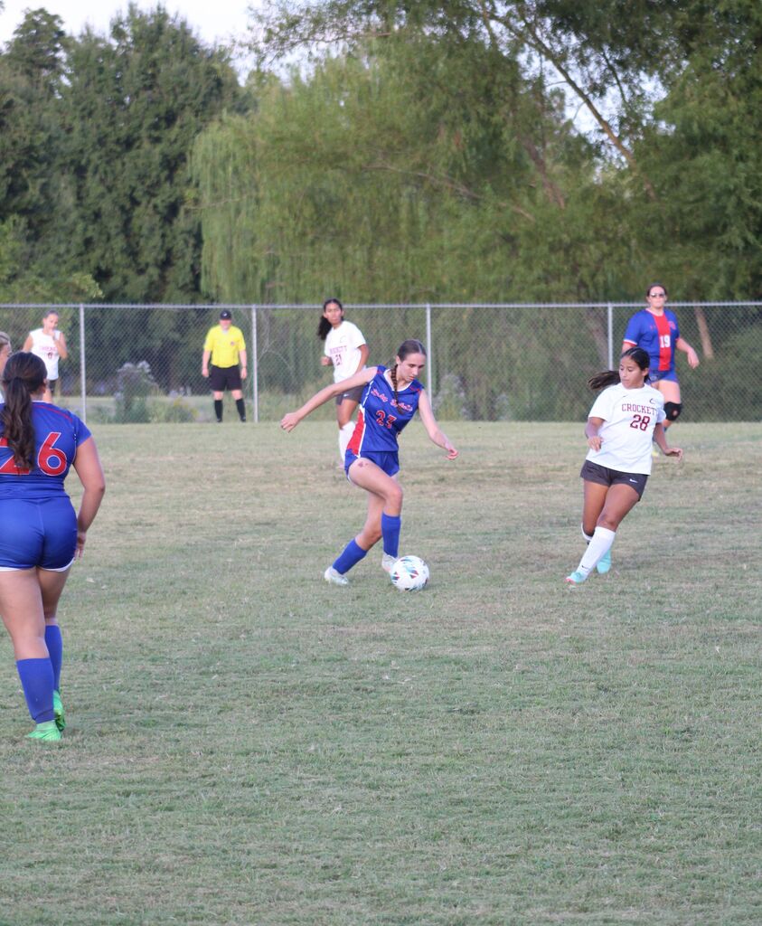 Girls soccer players in blue and white uniforms compete for the ball during a game on a grassy field, with teammates and a referee visible in the background.