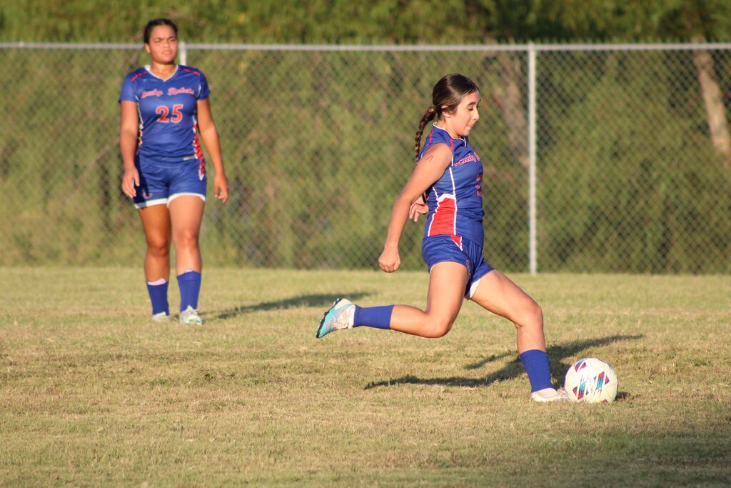 A girls soccer player in a blue uniform dribbles the ball across the field during a game, with a teammate in a matching uniform in the background.