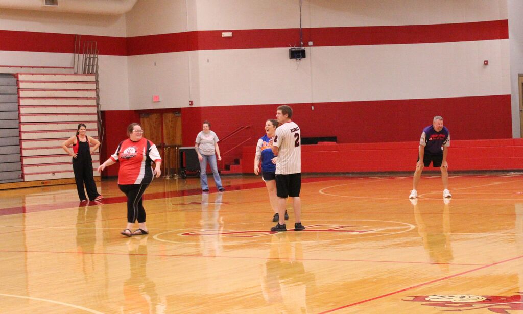 Several adults participate in a recreational game on an indoor gym floor, with red and white walls, bleachers, and a basketball court visible in the background.