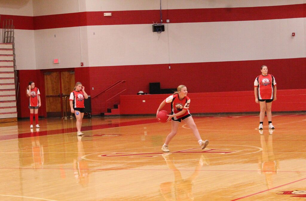 Four girls in red-and-black uniforms play a recreational game in a school gym. One girl in the center runs while holding a red ball, while three teammates stand nearby.