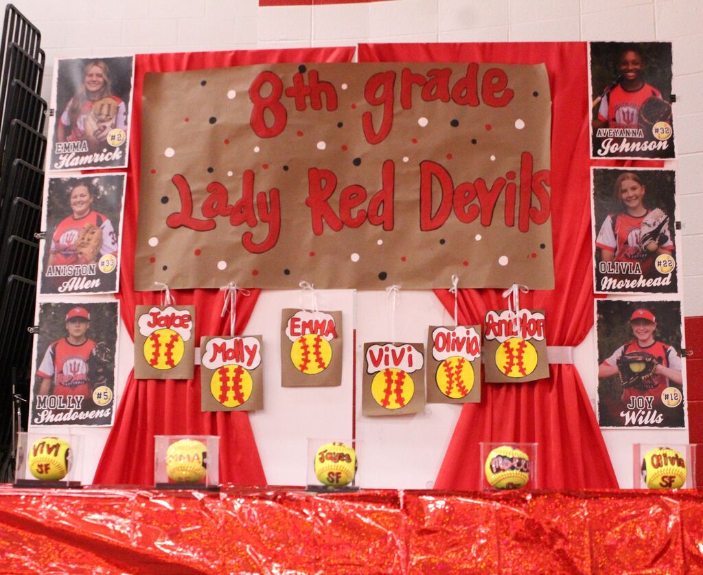 A red and white backdrop that holds a sign reading "8th Grade Lady Red Devils" with pictures of each 8th grade softball player.