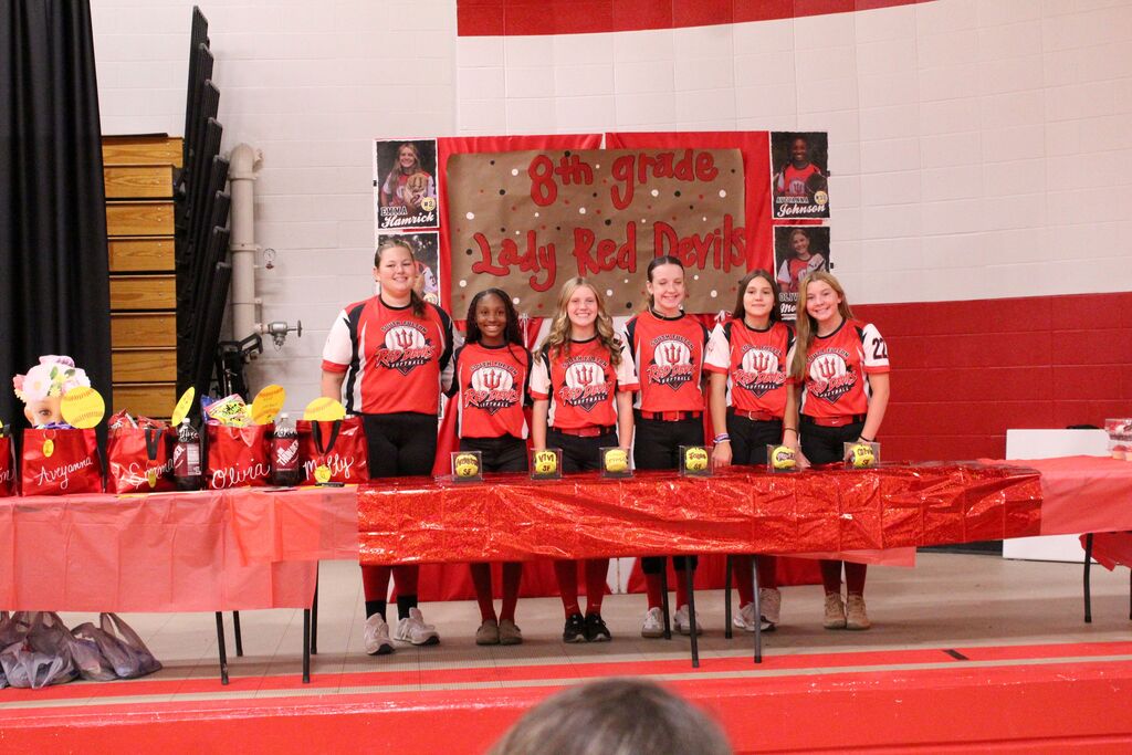 Six South Fulton Red Devils softball players in red-and-black uniforms stand behind a decorated table during an 8th grade celebration. A sign behind them reads “8th Grade Lady Red Devils,” with player photos displayed around it and softball-themed gifts on the tables.