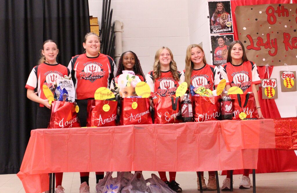 Six South Fulton Red Devils softball players stand behind a decorated table during their 8th grade celebration. Each player has a personalized red gift bag filled with softball-themed items, with an “8th Grade Lady Red Devils” display and player photos in the background.