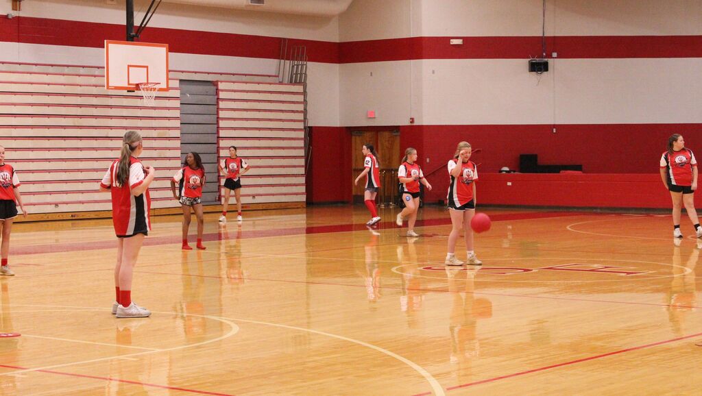 South Fulton softball players in red-and-black uniforms participate in a recreational game in a school gym, with one player holding a red ball near center court and teammates positioned around the court.