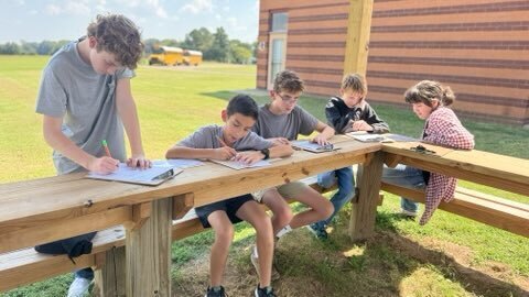 A group of OCMS students completes a class assignment together at outdoor wooden tables.