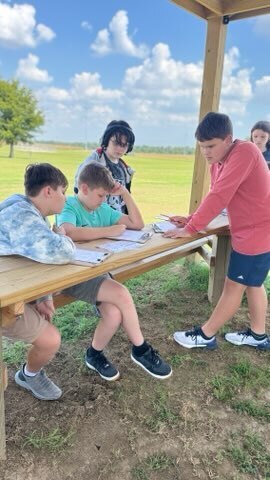A group of OCMS students completes a class assignment together at outdoor wooden tables.