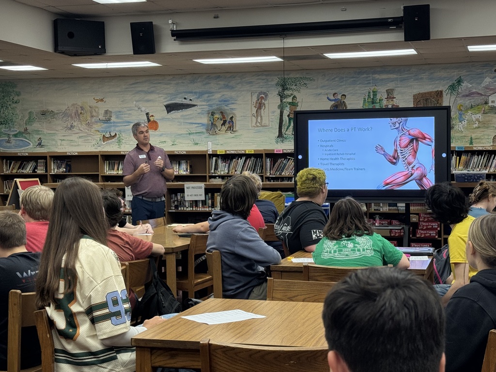 A guest speaker talks with OCMS students about careers in physical therapy while a presentation about where physical therapists work is displayed on the screen.