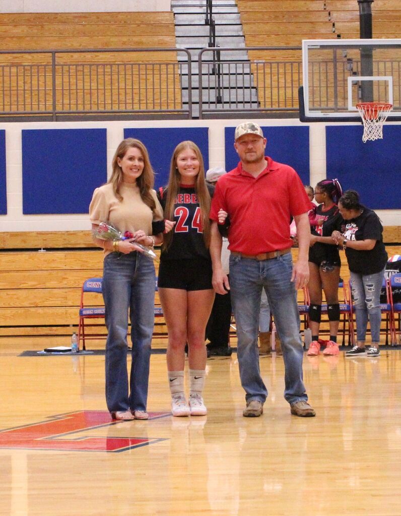 Three people stand together on a gymnasium basketball court during a senior recognition event. A volleyball player wearing a black “REBELS” jersey with the number 24 stands in the center between two adults. One adult holds a small bouquet of flowers. Wooden bleachers, a basketball hoop, blue wall padding, and folding chairs are visible in the background.