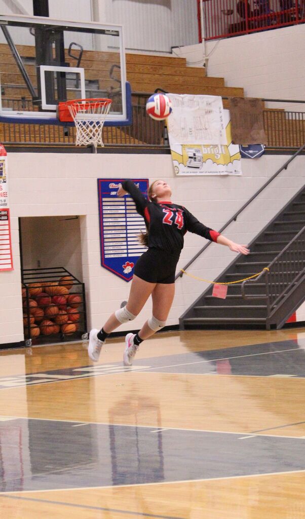 A volleyball player wearing jersey No. 24 leaps into the air during a game, reaching toward a red-and-white volleyball near the basketball goal. She is wearing a black uniform with red accents, black shorts, white knee pads, and athletic shoes. The gymnasium features wooden bleachers, a basketball hoop, and school banners in the background.