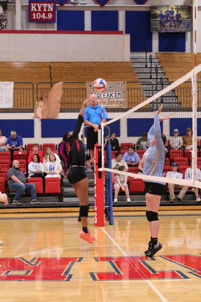 A volleyball player in a black and red uniform jumps near the net to hit the ball while an opposing player in a light blue uniform reaches up to block. A referee stands at the net, with spectators seated in red bleachers in the background.
