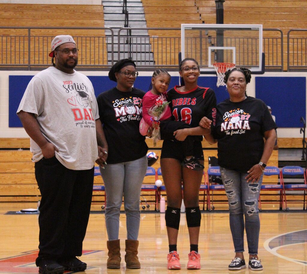 Group of five people standing on a high school gymnasium basketball court during a senior recognition event. One volleyball player wearing a black-and-red jersey with the number 16 stands in the center holding a young child and a bouquet of flowers. Three accompanying adults wear shirts with “Senior 27” designs, and basketball hoops, bleachers, and team chairs are visible in the background.