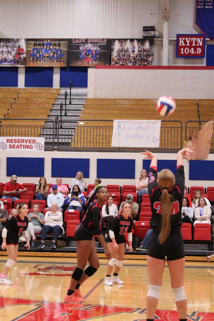 A volleyball player wearing No. 24 raises both arms to play the ball as teammates No. 12, No. 1, and another teammate watch the play develop. The red, white, and blue volleyball is airborne above the court. Spectators sit in red bleachers, with Lady Rebels volleyball team banners displayed along the gym wall.