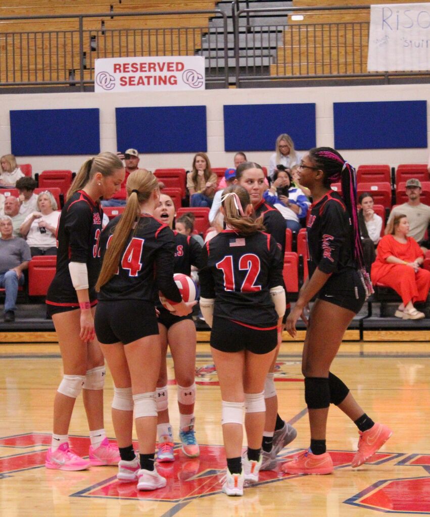 Several volleyball players in black and red uniforms gather on the court between plays. Spectators sit in red bleachers in the background.
