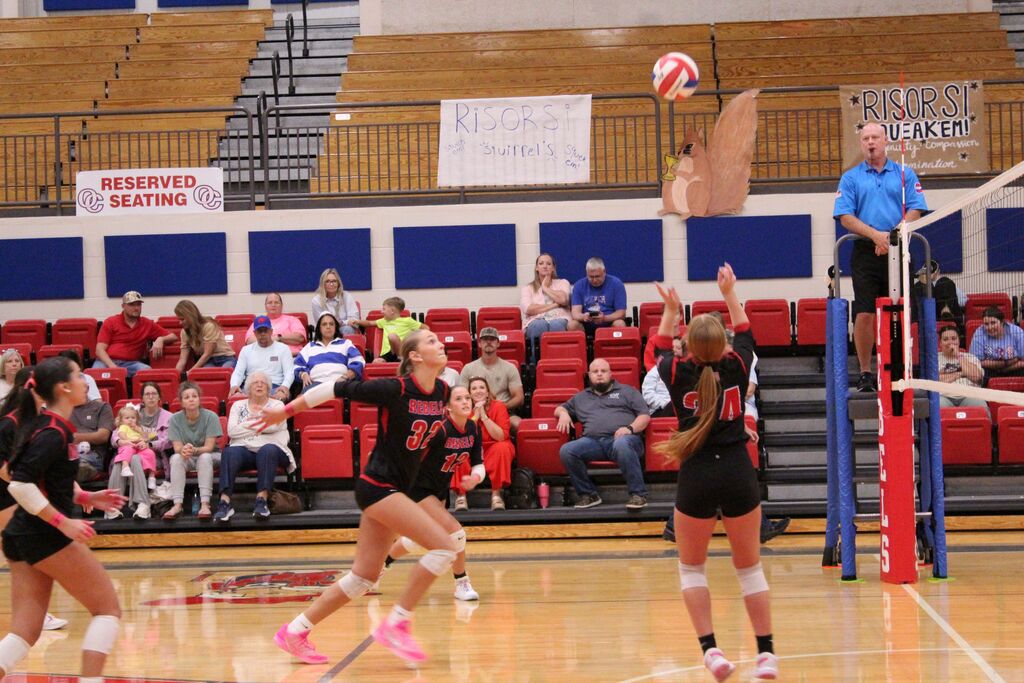 Volleyball players in black Rebels uniforms prepare for a play near the net during a match, with the ball in the air and spectators seated in the gymnasium bleachers behind them.