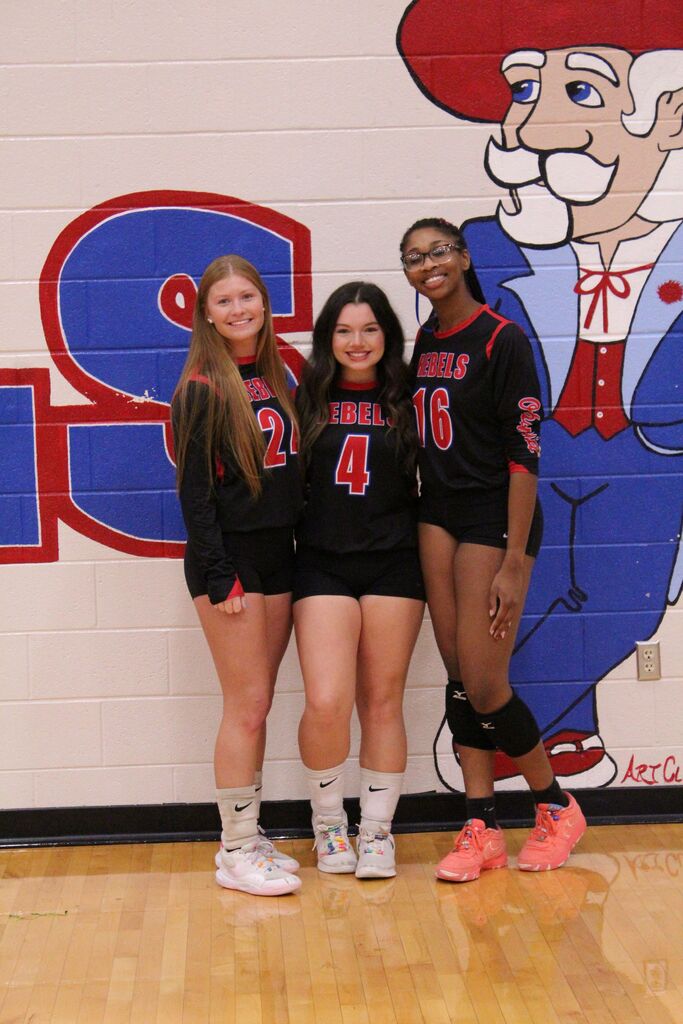 Three senior volleyball players wearing black and red Rebels uniforms pose together for a photo in front of a school mascot mural. The players wear jerseys No. 24, No. 4, and No. 16 and are smiling at the camera on the gymnasium floor.