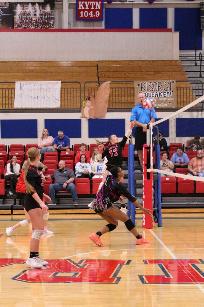 A volleyball player wearing No. 12 reaches up to hit the ball near the net as a teammate prepares behind her. Another teammate in a black and red uniform moves into position to make a play. The team plays on a gym floor with red and blue markings, with spectators seated in the bleachers behind them.